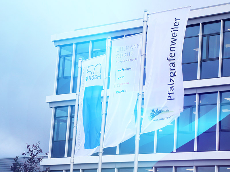 Three flags in front of a glass office building, showing 50 years of KOCH, the Uhlmann Group and Pfalzgrafenweiler